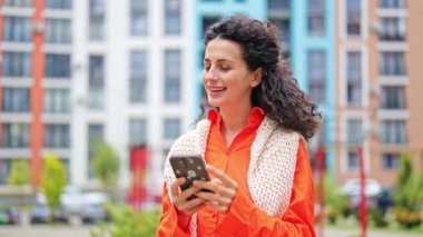 Turuncu gömlekli ve beyaz örgü şallı neşeli kadın telefonda iyi haberleri sever. Şık bir kadın konut binasının yanında mutlu olur. Modern mimari.