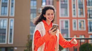 Neşeli, şık bir kadın açık havada parlak turuncu bir gömlek ve örgü bir yelek giyerek yürür. Mutlu bir şekilde göğsüne akıllı telefon tutarak başarısını kutlar..