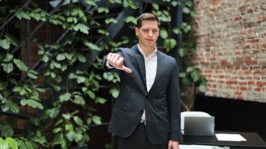 Takım elbiseli genç bir işadamı baş parmağını kaldırıyor. Modern ofiste yeşil bitkilerle yapılan jestlerden nefret ediyorum. Kötü geri bildirim, olumsuz duygu konsepti.