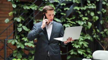 Kendine güvenen iş adamı dizüstü bilgisayar kullanıyor ve yeşil duvarın yanında telefonla konuşuyor. Modern ofiste çalışan ve pazarlık yapan profesyonel erkek girişimci..