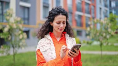 Modern binanın yakınında telefon kullanan neşeli genç bir kadın. Turuncu ve beyaz yazı yazan şık bir kadın internette geziniyor. Sakin ve olumlu duygular.