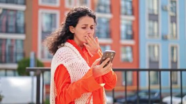 Şok olmuş turuncu elbiseli ve beyaz örgü yelekli bir kadın şehir caddesinde duruyor ve akıllı telefondaki mesajlara duygusal tepki veriyor..