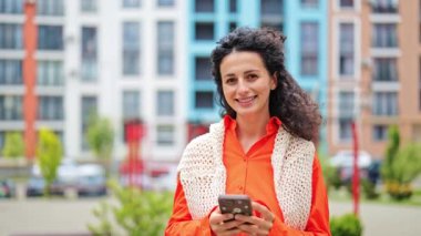 Turuncu tişörtlü, beyaz örülü yelek giyen güzel, neşeli bir kadın şehirdeki caddelerde mutlu bir şekilde telefonla gülüyor..