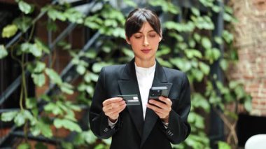 Telefon ve kredi kartı kullanan takım elbiseli neşeli bir iş kadını. Mutlu kadın çevrimiçi ödeme yapıyor. Ofis koridorunda başarılı yatırımcı.