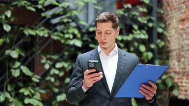 Takım elbiseli başarılı bir işadamı akıllı telefon kullanıyor ve elinde dosya tutuyor. İş adamı modern ofiste neşe hissediyor. Arkaplanda yeşil bitkiler.