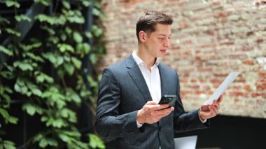Akıllı telefon kullanan başarılı bir işadamı modern ofiste belgeler ve gülümsemeler barındırır. Resmi bir takım giyer ve mutlu olur..