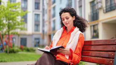 Dışarıda kitap okuyan güzel bir kadın. Bayan öğrenci temiz havada ilginç bir hikayenin tadını çıkarıyor. Turuncu gömlek ve beyaz yelek giyen kıvırcık esmer.