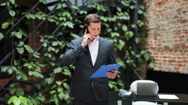 Başarılı bir iş adamı telefonda konuşuyor ve belgeleri tutuyor. Modern ofiste çalışan takım elbiseli yakışıklı adam. Kapalı, iş, iş.