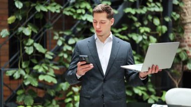 Takım elbiseli neşeli iş adamı akıllı telefon ve dizüstü bilgisayar kullanıyor modern ofiste kameraya gülümsüyor. İş ve teknoloji kavramı.