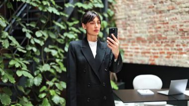 Modern ofiste akıllı telefon kullanarak şık giyinen kendine güvenen bir iş kadını. Yeşil bitkiler ve tuğla duvarlar rahatlatıcı bir ortam yaratır. Video çağrısı ile iletişim kuruyor.