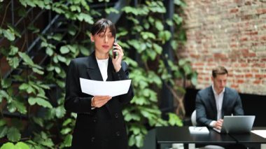 Resmi giyinmiş kendine güvenen bir iş kadını iş meselelerini telefonda tartışıyor, yeşil ofis alanındaki belgeleri gözden geçiriyor. Adam arka planda çalışıyor.