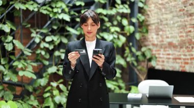 Siyah takım elbiseli neşeli iş kadını telefon ve kredi kartıyla online ödeme yapıyor. Yeşil duvarlı modern ofis alanı. Tatmin, mutlu, başarılı..