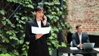 Resmi giyinmiş kendine güvenen bir iş kadını modern ofisteki cep telefonu görüşmelerinde dokümanları tartışır. İş adamı arka planda çalışır.