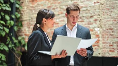 Resmi giyimde başarılı iş ortakları, bitkilerle tuğla bir duvara yaslanmış dizüstü bilgisayar ve belge kullanarak bir projeyi tartışıyorlar..