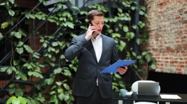 Takım elbiseli kendine güvenen bir işadamı telefonda konuşuyor ve modern ofiste dokümanları okuyor. Proje detaylarını tartışan profesyonel bir işçi. Başarılı bir adam telefonla arıyor..