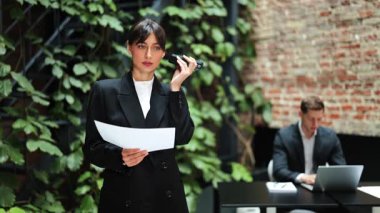 Siyah takım elbiseli kendine güvenen bir iş kadını modern bir ofiste sesli mesaj yazdırıyor. İş kadını yeşil bitkilerle dolu bir duvarın yanında duruyor..