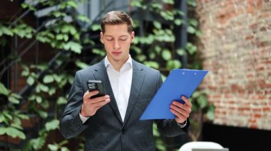 Takım elbiseli neşeli iş adamı telefon kullanıyor ve klasörü tutuyor. Modern ofiste kameraya gülümsüyor. Mutlu işçi, başarılı iş, yeni teknolojiler.