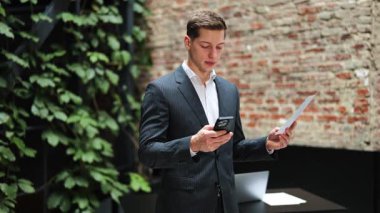 Telefon kullanarak ve belgeleri analiz ederek takım elbiseli bir iş adamı. Duvarları ve bitkileri olan modern bir ofis. Profesyonel iş, konsantrasyon ve iş stratejisi.