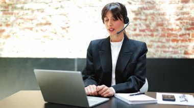 Takım elbiseli ve kulaklıklı kendine güvenen bir iş kadını laptopta çalışıyor ve ofisteki çevrimiçi bir toplantıda konuşuyor. Coşkulu hissediyor..