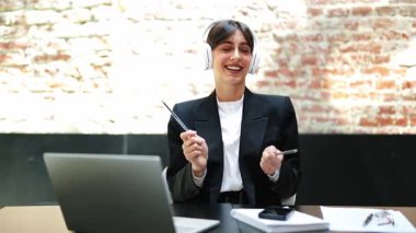 Resmi takım elbiseli neşeli iş kadını müzik dinler ve kalemlerle dans eder. Ofis katında tuğladan duvarlarla eğlenir..