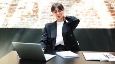 Takım elbiseli yorgun bir iş kadını ofiste dizüstü bilgisayarla çalışırken boynuna masaj yapıyor. Acı ve stres hissediyor..