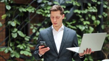 Kendine güvenen takım elbiseli bir işadamı elinde dizüstü bilgisayarla yeşil duvarın yanında duruyor. Modern ofis, iş, teknoloji, başarılı adam.