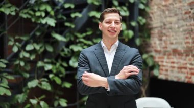 Takım elbiseli neşeli iş adamı modern ofiste kollarını kavuşturup gülümsüyor ve mutlu ve başarılı hissediyor. Arka planda yeşil bitkiler ve tuğla duvar.