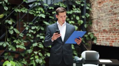 Akıllı telefon kullanan ve modern ofiste pano tutan kendine güvenen bir iş adamı. Kendini odaklanmış ve üretken hissediyor. İş günü resmi takım elbiseli şık bir işadamı..