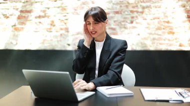 Ofiste baş ağrısı çeken takım elbiseli stresli bir iş kadını. O bir masada oturuyor ve bir dizüstü bilgisayar üzerinde çalışıyor. Ofiste tuğla duvar var..