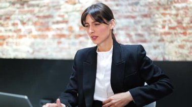 Resmi giyinmiş genç bir iş kadını güçlü bir mide ağrısı çekiyor. Modern ofiste tuğla duvarlı bir iş kadını oturuyor. Kadın acı çekiyor ve karnını tutuyor..