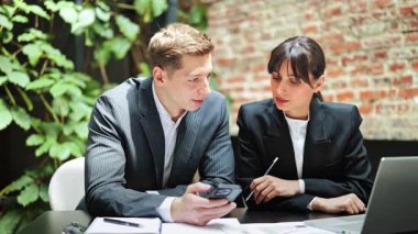 Resmi olarak kendine güvenen iş arkadaşları modern ofis alanında proje detaylarını tartışırken dizüstü bilgisayar ve telefon kullanıyorlar, odaklanmış ve kararlılar..