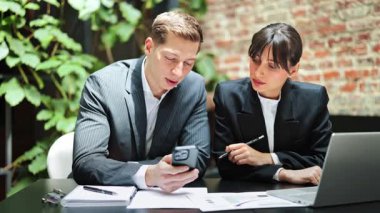 Resmi giyimde odaklanmış meslektaşlarım modern bir ofiste bir projeyi tartışıyorlar. Bir telefon ve dizüstü bilgisayardaki verileri analiz ederler, takım çalışması ve konsantrasyonu gösterirler..