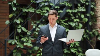 Akıllı telefon kullanan ve modern ofiste dizüstü bilgisayar tutan odaklı bir iş adamı. Zarif takım elbiseli başarılı bir adam. İş ve teknoloji kavramı.