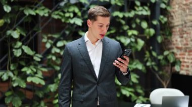 Heyecanlı takım elbiseli iş adamı telefonu kullanıyor, mutlu hissediyor. Başarılı yönetici zaferi bitkilerle kutluyor. Neşe, şans.