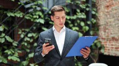 Takım elbiseli kendine güvenen iş adamı akıllı telefon kullanıyor ve ofis yeşilliğinin arka planında pano duruyor. Gülümsüyor ve mutlu görünüyor..