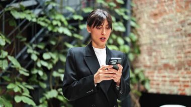 Modern bir ofiste telefon kullanan siyah takım elbiseli ve beyaz gömlekli profesyonel bir iş kadını. Şaşırmış ve biraz endişeli hissediyor..