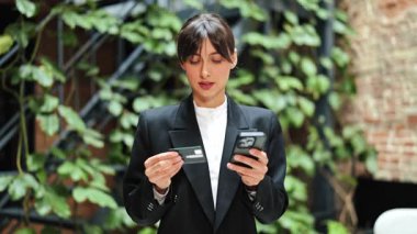 Yeşil duvarın yanında akıllı telefon ve kredi kartı kullanan resmi giyinmiş kendine güvenen bir iş kadını. Modern ofiste kolay çevrimiçi ödeme.