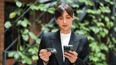 Telefon ve kredi kartı kullanan takım elbiseli neşeli bir iş kadını. Çevrimiçi ödeme yapan mutlu bir kadın, açık havada başarılı bir e-ticaret..