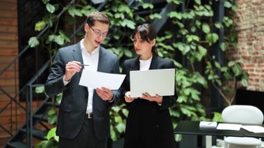 Resmi kıyafet giyen profesyonel meslekdaşlar bir ofiste fabrikaların yanında durur ve belge ve dizüstü bilgisayar kullanarak bir projeyi tartışırlar..