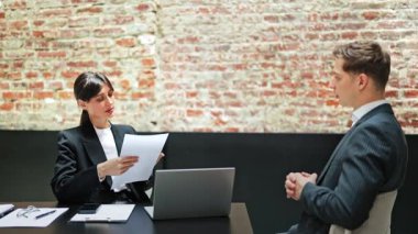 Takım elbiseli kendine güvenen bir iş kadını, bir işadamıyla strateji tartışan belgeler tutuyor. Ofiste tuğla duvarlı bir iş toplantısı..