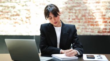 Modern ofiste çalışan resmi giyinmiş kendine güvenen bir iş kadını. Deftere yazıyor, dizüstü bilgisayar kullanıyor ve kameraya gülümsüyor..