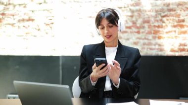 Resmi giyinmiş neşeli iş kadını çalışırken müziğin keyfini çıkarıyor. Modern ofisinde ritme ayak uydururken gülümsüyor..