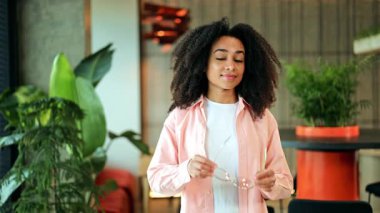 Güzel bir Afrikalı Amerikalı kadın şık gözlük takar ve kendine güvenerek modern bir ofiste gülümser. Beyaz tişört ve pembe gömlek giyen serbest çalışan, pozitif ve neşeli duygular yayıyor..