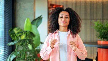 Pembe tişört ve beyaz tişört giyen neşeli, profesyonel Afro-Amerikan kadın, modern ofis ortamında güven içinde poz vererek gözlüğünü mutlulukla çıkarıyor. Pozitif enerjisi neşe saçıyor..