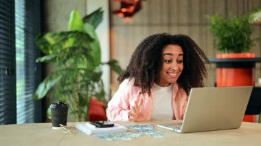 Heyecanlı, beyaz tişörtlü, pembe tişörtlü Afrikalı bir kadın modern ofis masasında finansal başarısını başarıyla kutluyor..