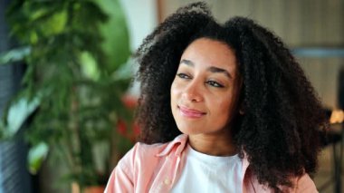 Pembe tişörtlü, kıvırcık saçlı, güzel Afro-Amerikan kadın kapalı alanda düşünceli ve huzurlu görünüyor. Kadın düşünceli, huzurlu gülümsemeler, sakince, sakin anları yansıtıyor..