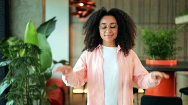 Pembe tişört ve beyaz tişört giymiş kıvırcık saçlı ve gözlüklü, kendine güvenen Afro-Amerikan kadın, bitkilerin arasında neşeli bir şekilde poz veriyor. Profesyonel ve olumlu bir duygu..