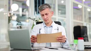 Beyaz önlüklü ciddi bir doktor modern ofiste tıbbi dokümanlarla ilgili bir şeyler okuyor. Deneyimli erkek doktor önemli belgeleri inceledikten sonra endişe ve stres ifade ediyor.