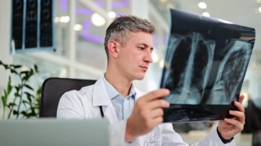 Profesyonel doktor modern bir muayenehanede oturup hastanın göğüs röntgenini inceliyor. Tıbbi uzman doğru teşhis için radyografiyi titizlikle analiz eder..