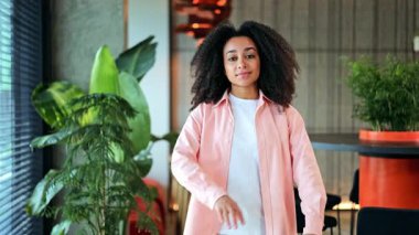 Kıvırcık saçlı güzel bir Afro-Amerikan iş kadını modern bir ofiste kendine güveniyor. Pembe bir gömlek giyer, gülümser, mesleki ortamında başarı ve mutluluk saçar..